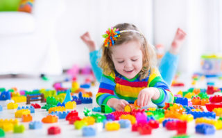 Cute funny preschooler little girl in a colorful shirt playing with construction toy blocks building a tower in a sunny kindergarten room